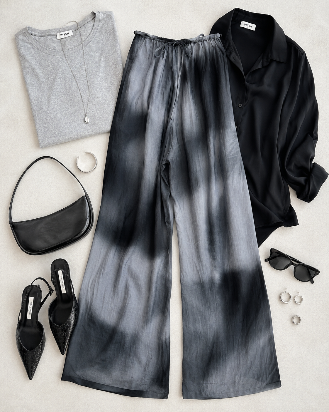 Flat lay of DISSH black and gray tie-dye wide-leg pants styled with a gray tee, black satin blouse, pointed shoes, handbag, and silver accessories.
